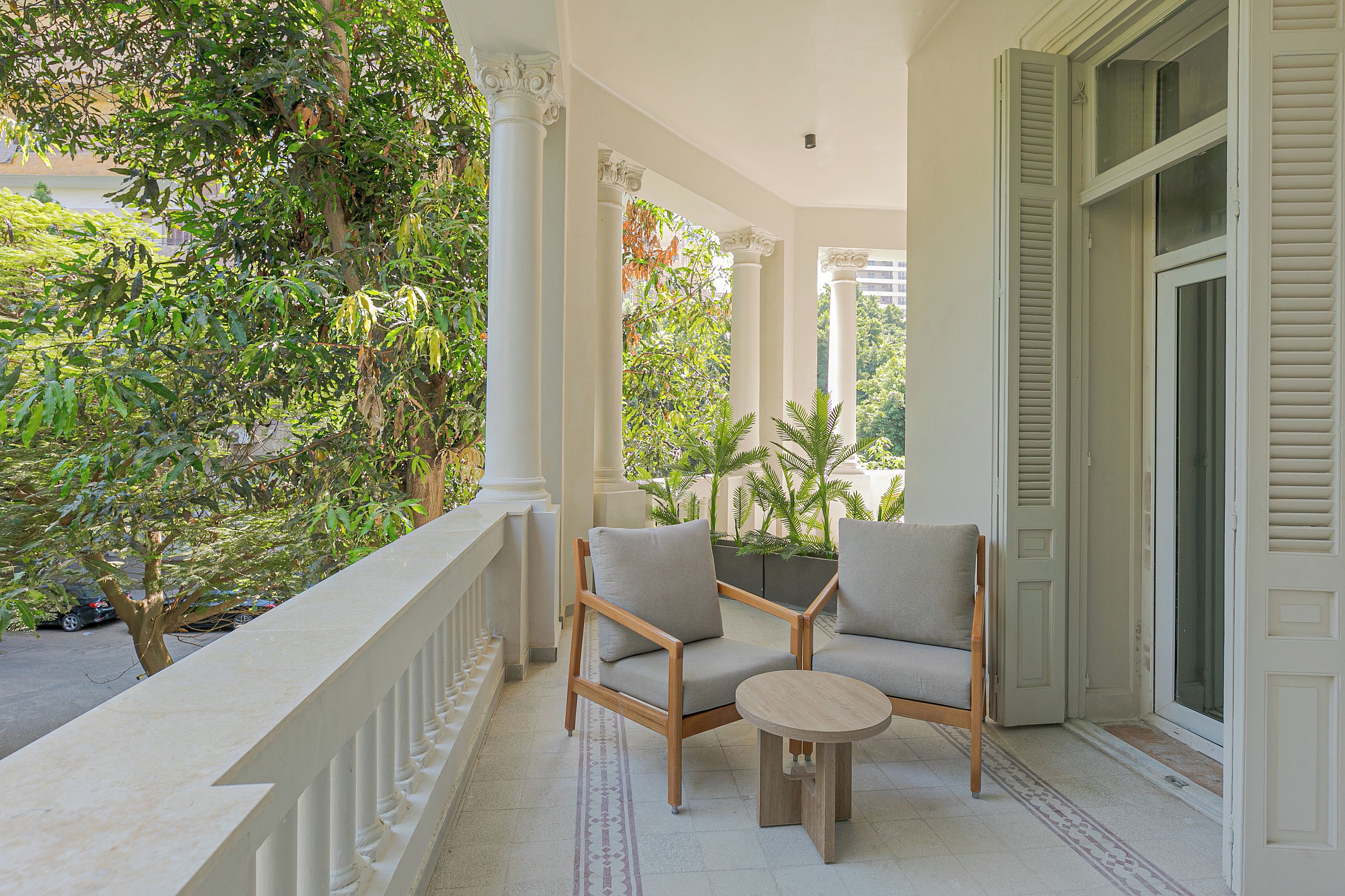 A covered outdoor seating area with two chairs and a small table, surrounded by white columns and a railing.