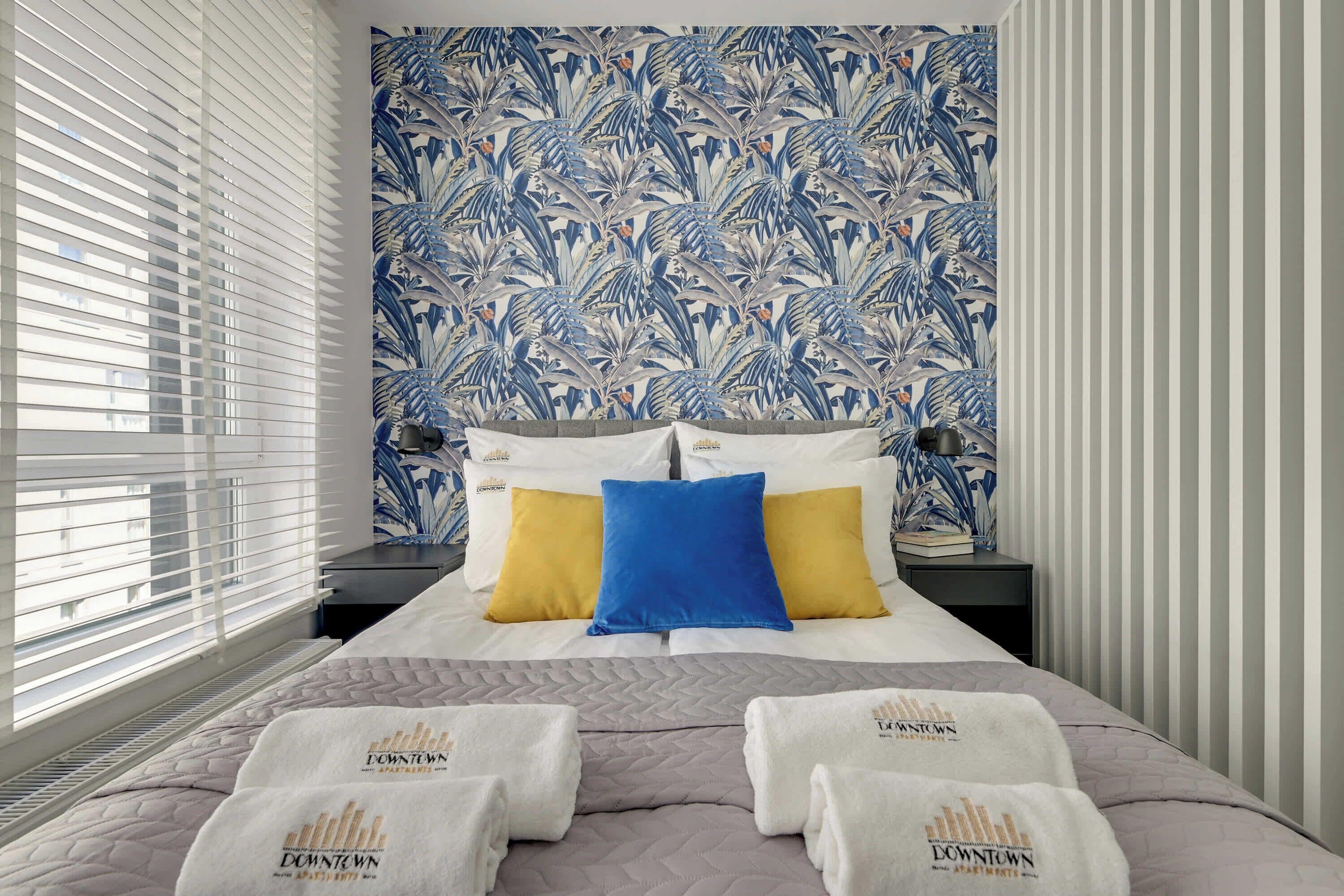 A hotel room with a bed, pillows, a nightstand, and a window with blinds.