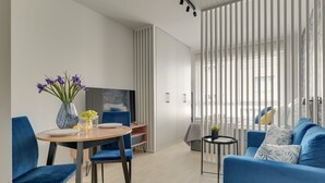 A modern living room with a blue sofa, a dining table with chairs, a TV, and a large window with blinds. - Śląska 307 | Gym | Perfect for Couples (Gdańsk)
