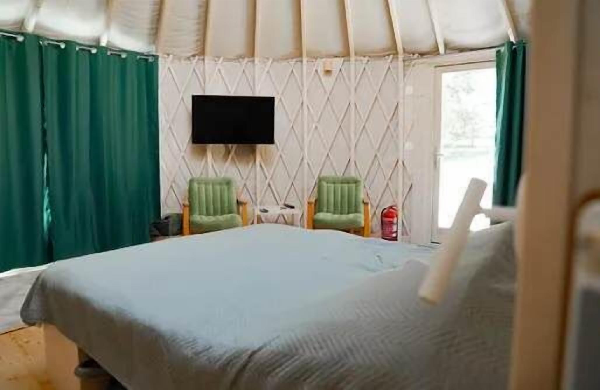 Ein Zelttyp-Zimmer mit einem Bett, zwei Stühlen, einem an der Wand montierten Flachbildfernseher und einem Fenster mit Vorhängen.