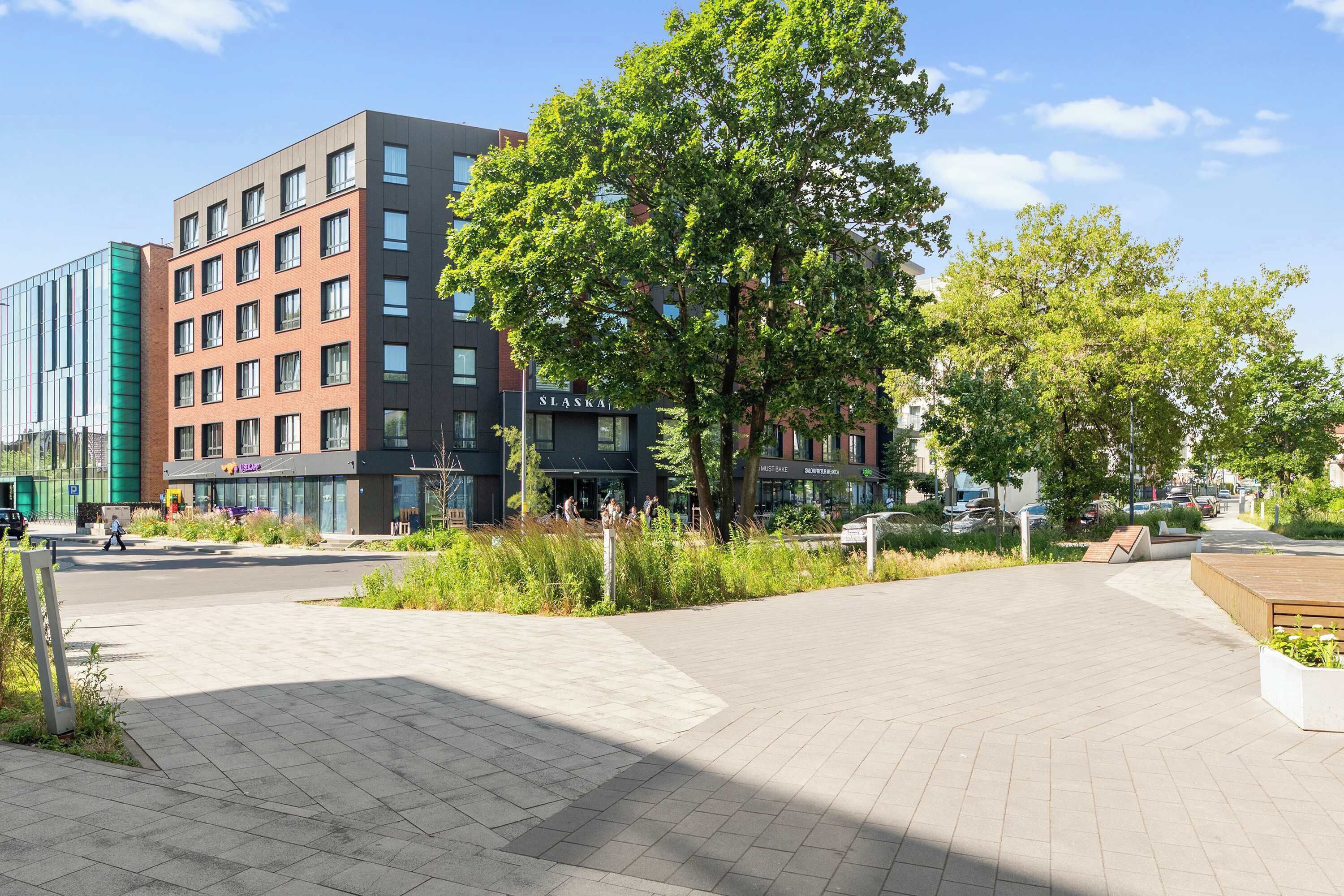 A modern building with a sign 'SLASKA' and a paved plaza with trees and benches.