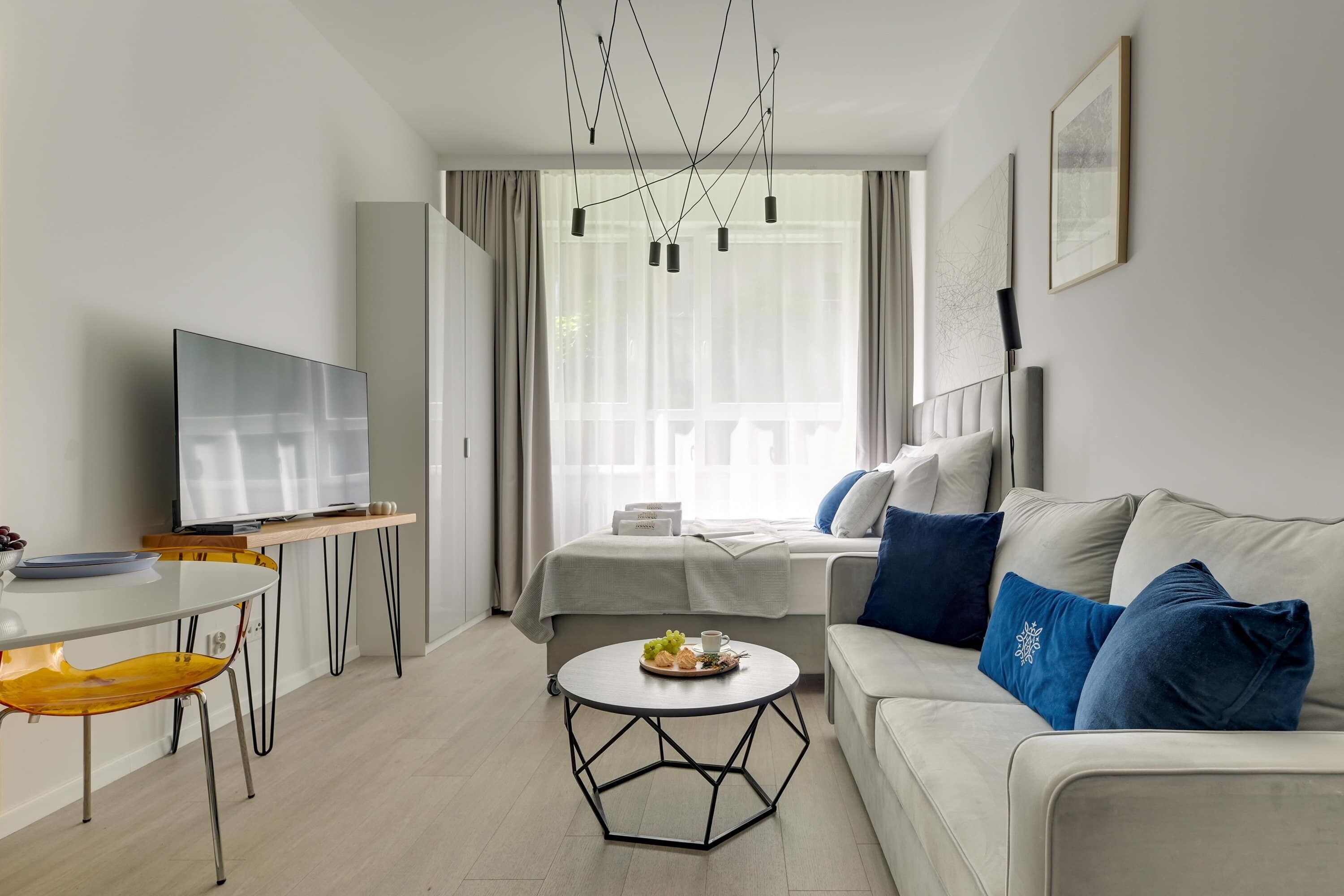 Une chambre d’hôtel moderne équipée d’un téléviseur à écran plat, d’une table à manger avec des chaises, d’un canapé avec des coussins bleus, d’une table basse ronde et d’un lit avec une couette blanche.