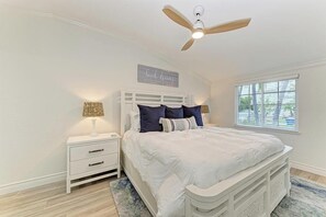 Quarto com cama, mesas de cabeceira, ventoinha no teto, janela com persianas e uma placa fixada na parede.