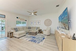 Uma sala de estar com um sofá seccional, uma mesa de centro, uma mesinha lateral, um abajur, um ventilador de teto e um quadro na parede.