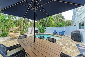 Um quintal com mesa e cadeiras de madeira, piscina e área de churrasco coberta.