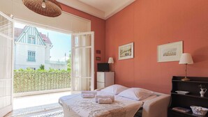 A bedroom with a bed, a desk, a chair, a lamp, and a window with a view of a building outside. - Flat in a Traditional Norman Townhouse - Garden (Deauville)