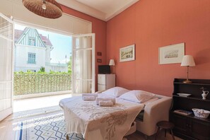 A bedroom with a bed, a desk, a chair, a lamp, and a window with a view of a building outside. - Flat in a Traditional Norman Townhouse - Garden (Deauville)