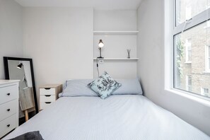Ein Schlafzimmer mit Bett, Nachttisch, Lampe und einem Fenster mit Blick auf ein Gebäude.