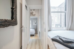 En smal gang der fører til et soveværelse med en seng, et vindue og en dør.