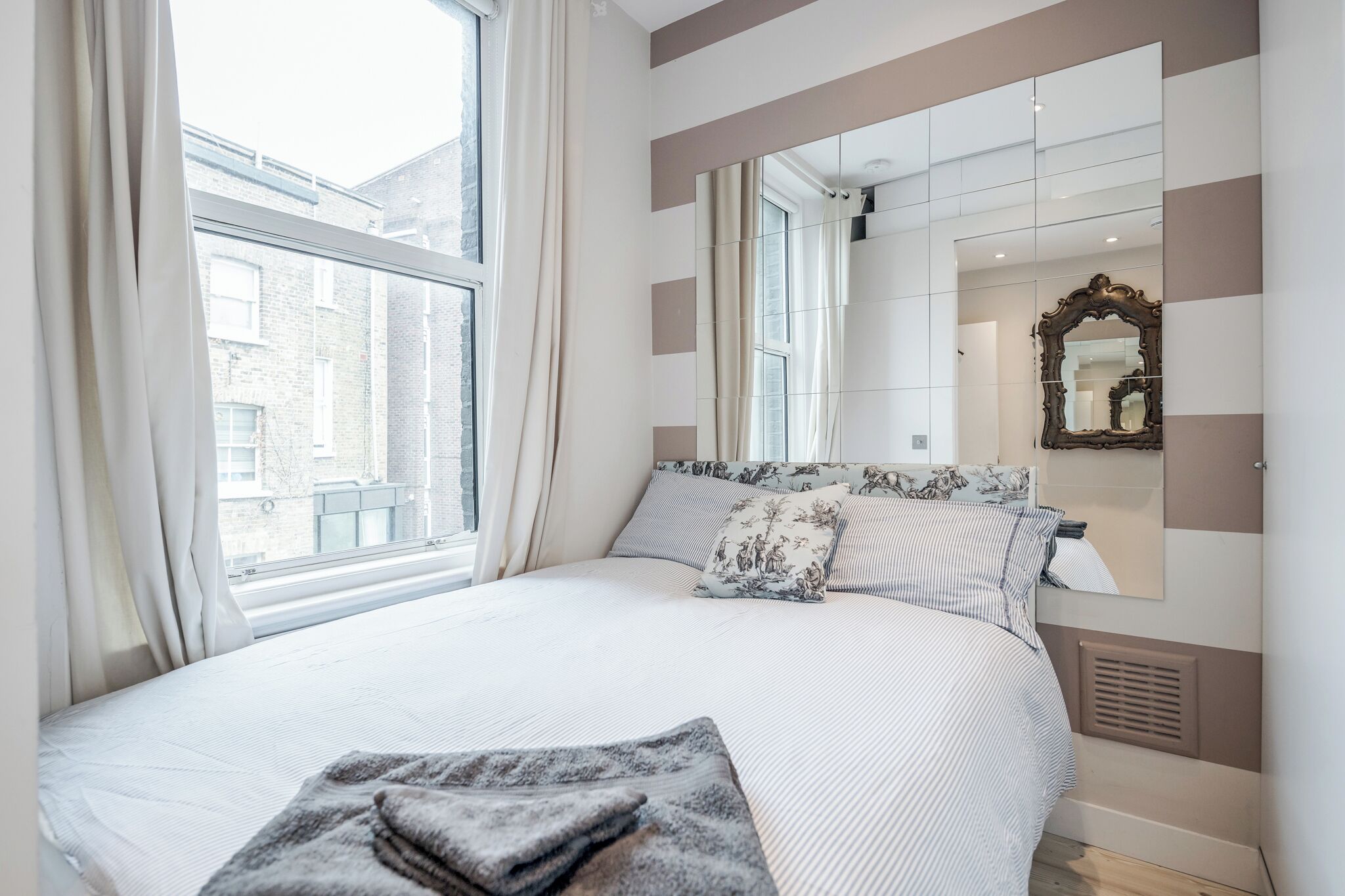A modern bedroom with a large bed, a window with curtains, a mirror, and a view of buildings outside.