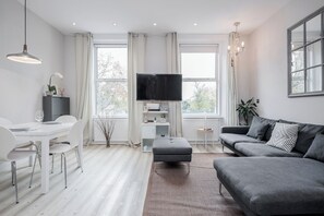 Ein modernes Wohnzimmer mit einem grauen Sofa, einem Esstisch mit Stühlen, einem Fernseher und einem großen Fenster mit Vorhängen.