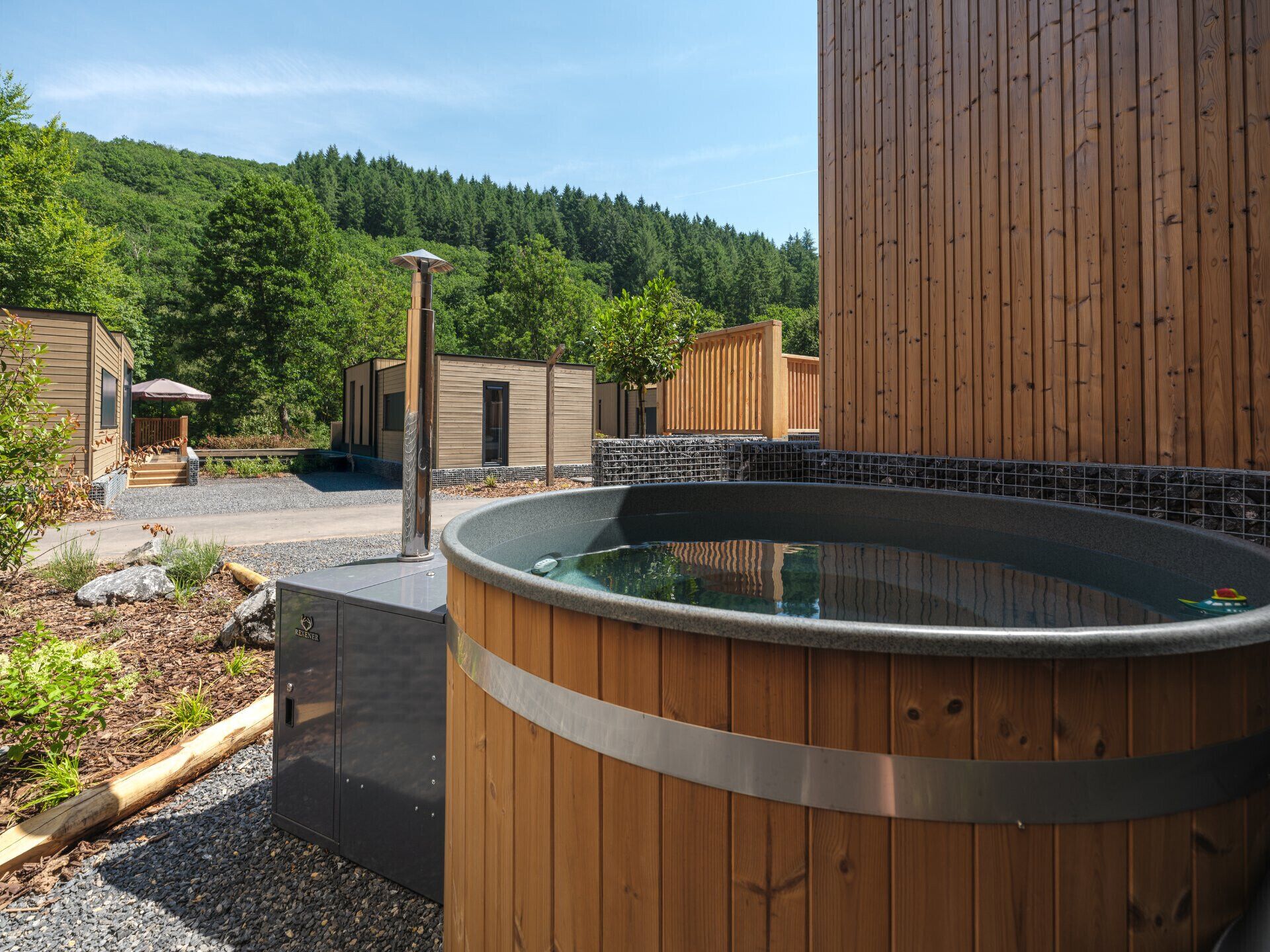 Un bain à remous entouré de structures en bois, un chemin de gravier et, au loin, une colline boisée.