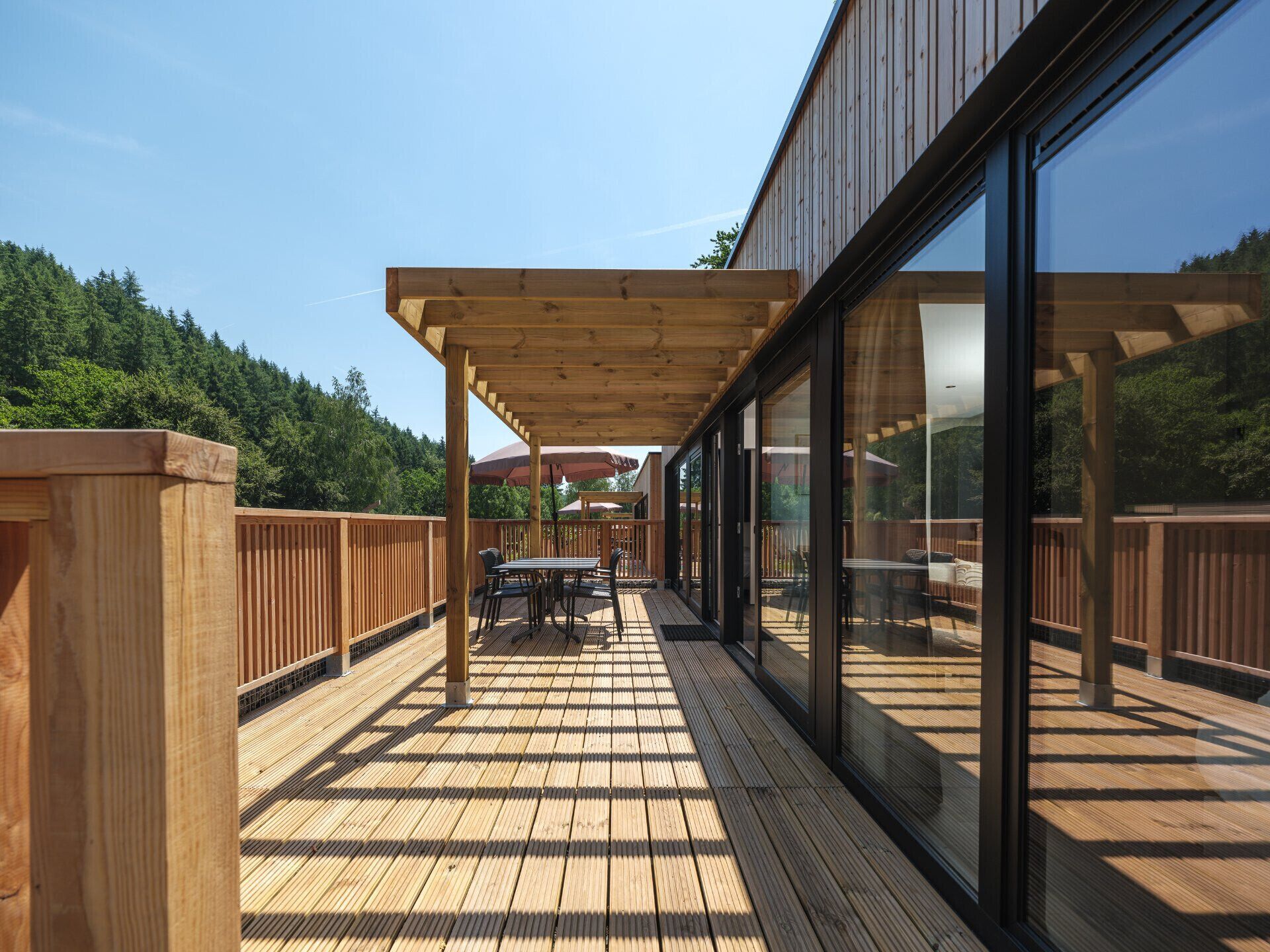 Une terrasse en bois agrémentée d’une pergola, de mobilier d’extérieur et de murs en verre offrant une vue sur la forêt.