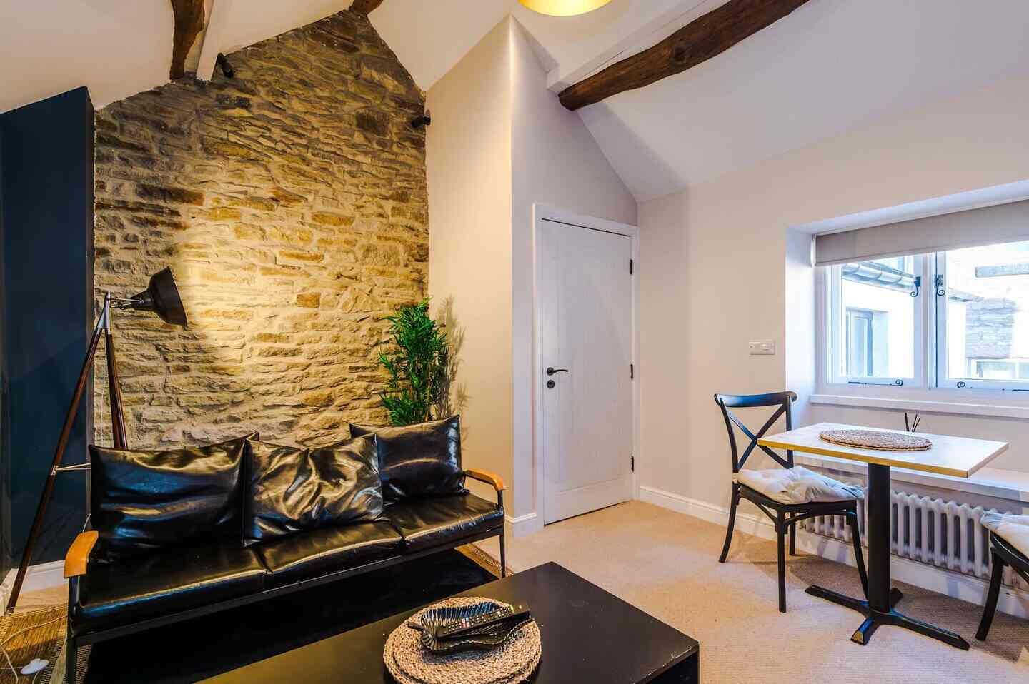 A cozy room with a stone wall, a black leather sofa, a dining table with chairs, and a window providing natural light.