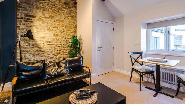 A cozy room with a stone wall, a black leather sofa, a dining table with chairs, and a window providing natural light. - The Vault - Boutique Cosy Peak District Retreat in a Grade II listed building (Chapel-en-le-Frith)