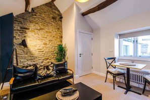 A cozy room with a stone wall, a black leather sofa, a dining table with chairs, and a window providing natural light. - The Vault - Boutique Cosy Peak District Retreat in a Grade II listed building (Chapel-en-le-Frith)