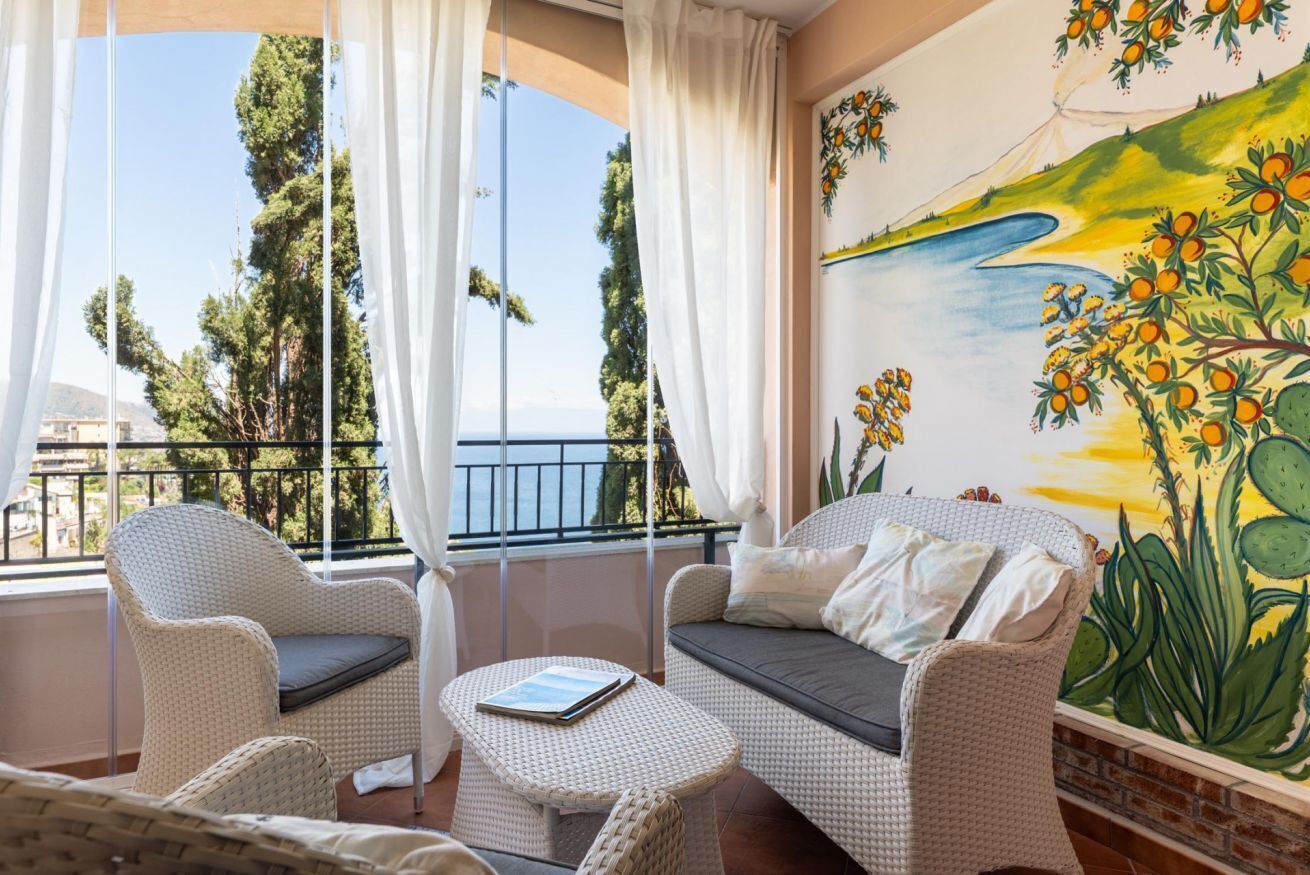 Een balkon met rieten meubilair, uitzicht op zee, en een muurschildering met cactussen en bloemen.
