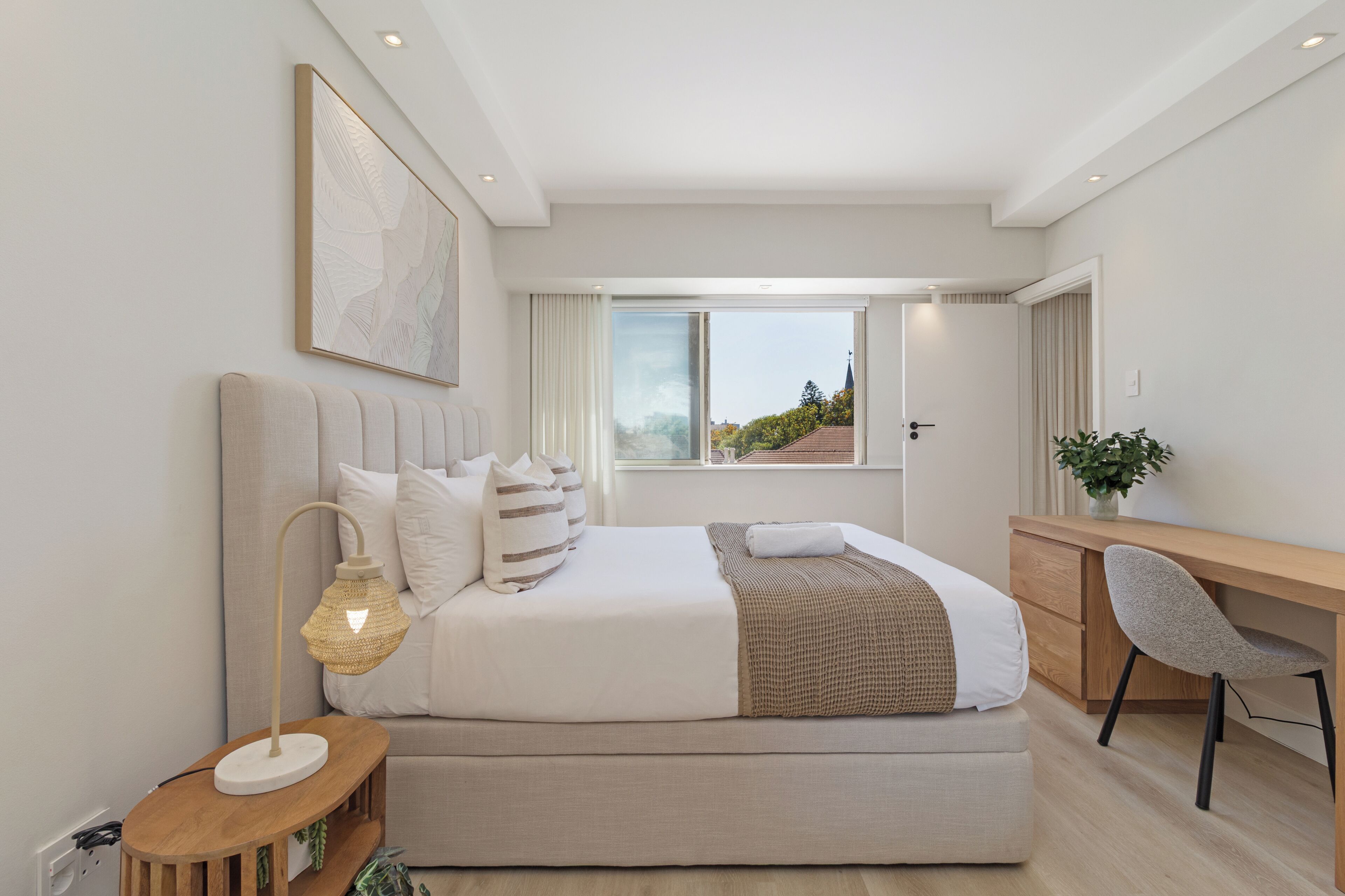 A modern bedroom with a large bed, bedside table, lamp, and a view of the outside.
