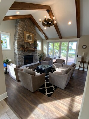 A spacious living room with a stone fireplace, wooden beams, and a large window offering a view of the outdoors.