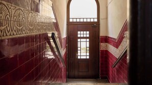 Un escalier avec une porte en bois et une fenêtre, menant à une pièce aux murs recouverts de carreaux rouges et ornée d’une rampe en métal décorative.