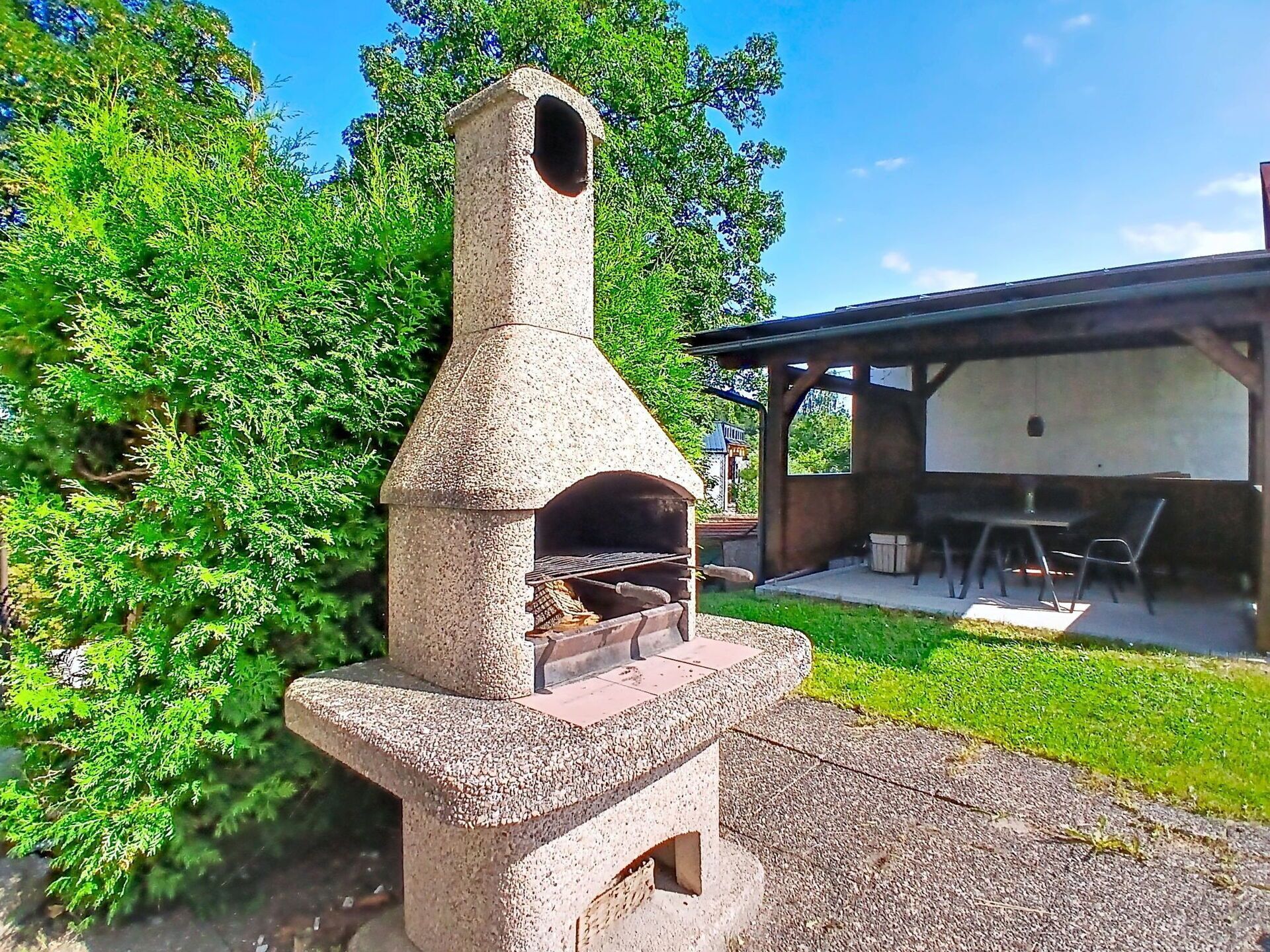 A stone-built outdoor pizza oven with a chimney, surrounded by greenery and a covered seating area.