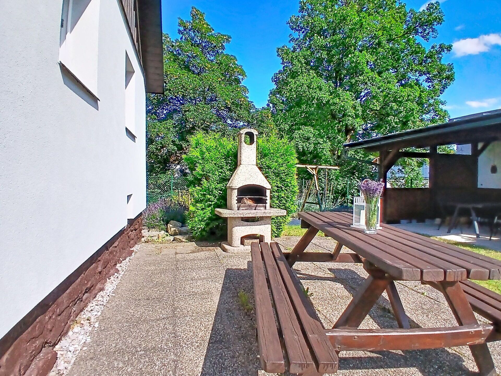 Een terras met een pizzaoven van natuursteen, een houten picknicktafel en een wit gebouw op de achtergrond.