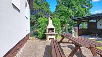 Een terras met een pizzaoven van natuursteen, een houten picknicktafel en een wit gebouw op de achtergrond.