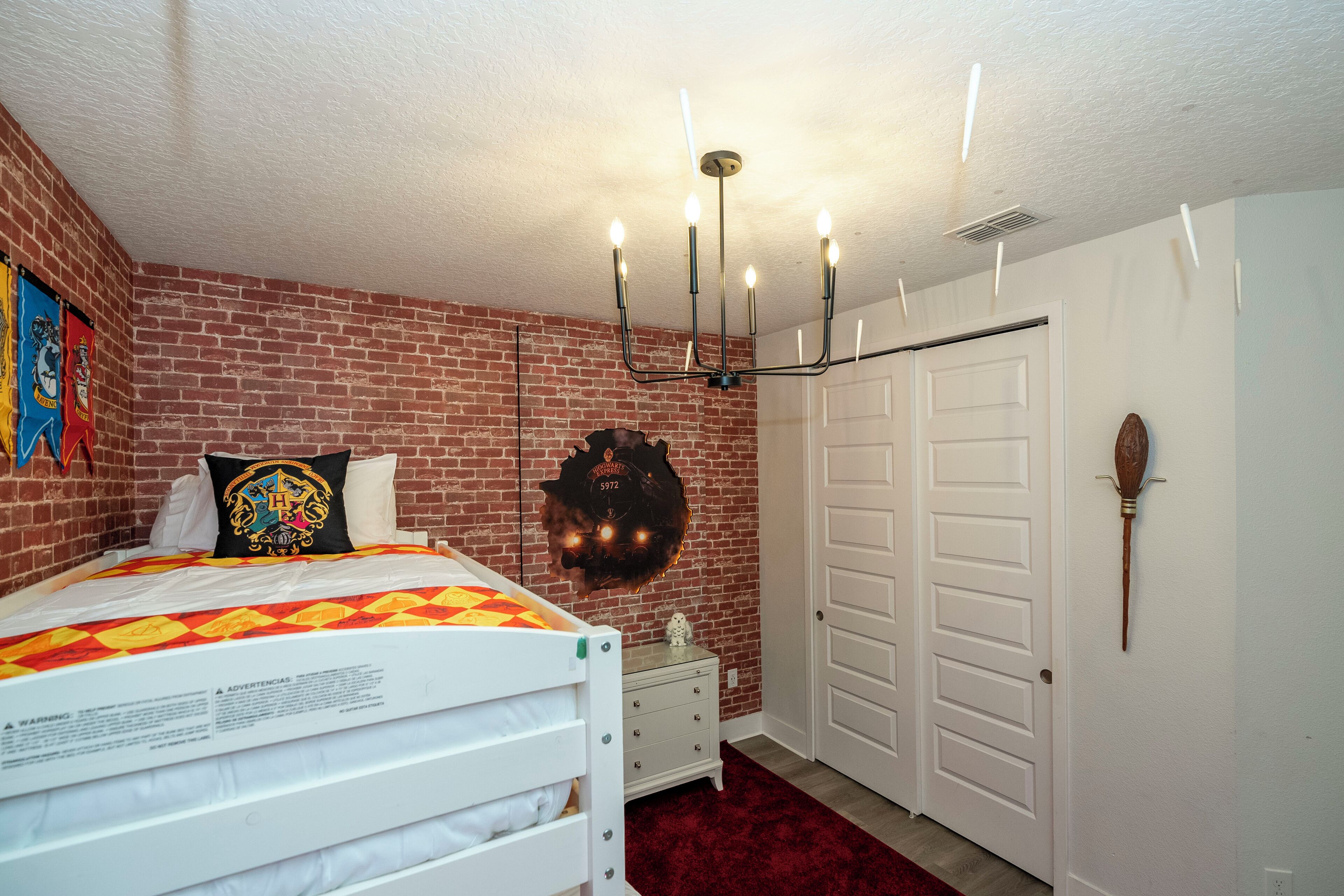 A bedroom with a brick wall, a bed with a patterned bedspread, a wall-mounted light fixture, and a door with a wand.
