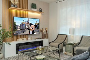 A modern living room with a flat-screen TV mounted on a wooden wall, a glass coffee table, two patterned armchairs, a side table with a lamp, and a fireplace with a blue-lit glass front. - Luxury Disney Escape - Pool, SPA and Games (Kissimmee)