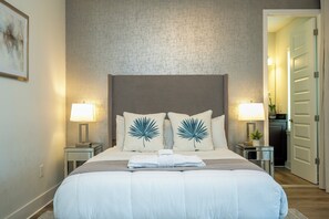 A hotel room with a large bed, two bedside lamps, a nightstand with a plant, and a door leading to another room. - Luxury Disney Escape - Pool, SPA and Games (Kissimmee)