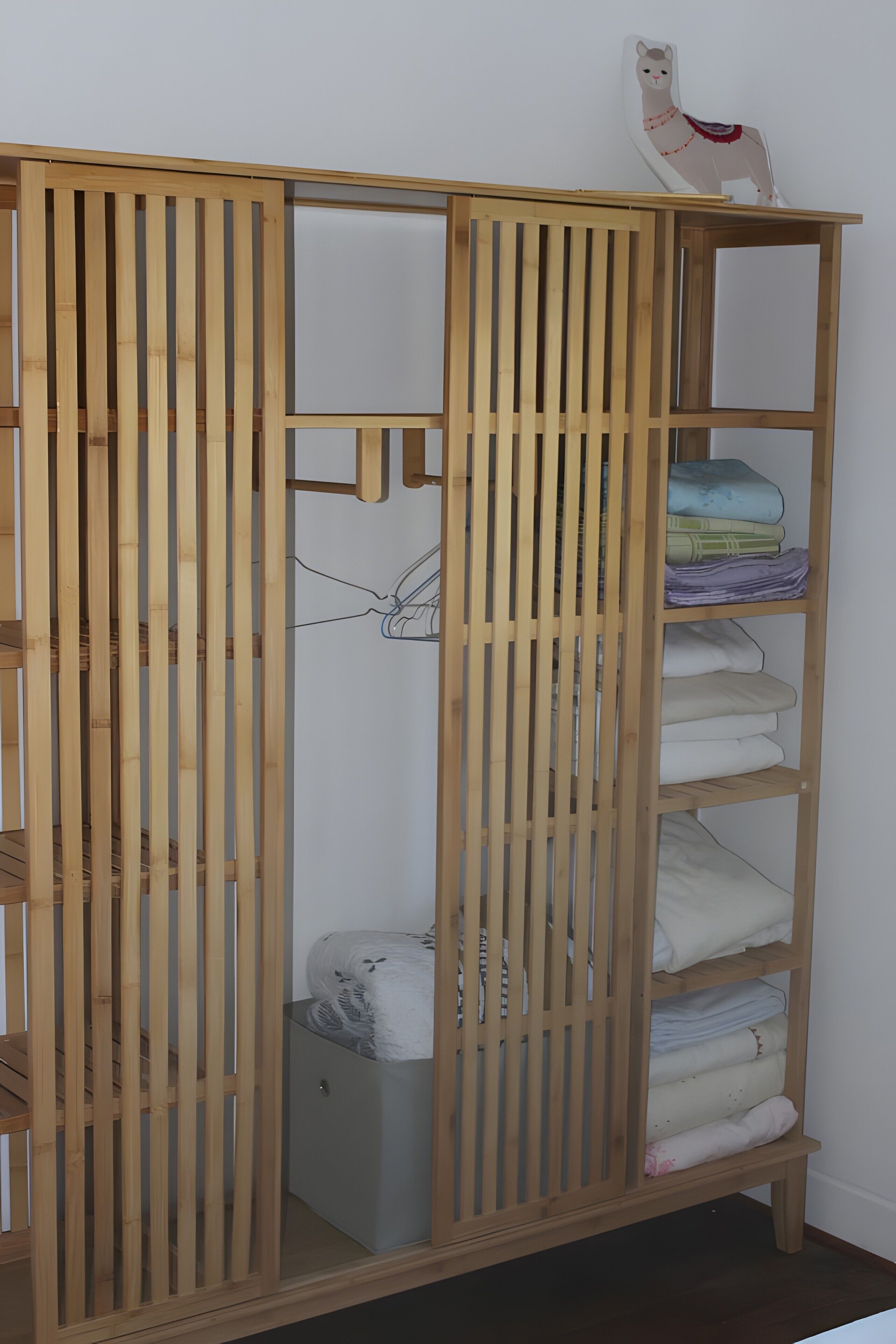 Une armoire en bois, dotée de plusieurs étagères et d’une tringle à vêtements, contenant des vêtements pliés et une petite boîte de rangement.