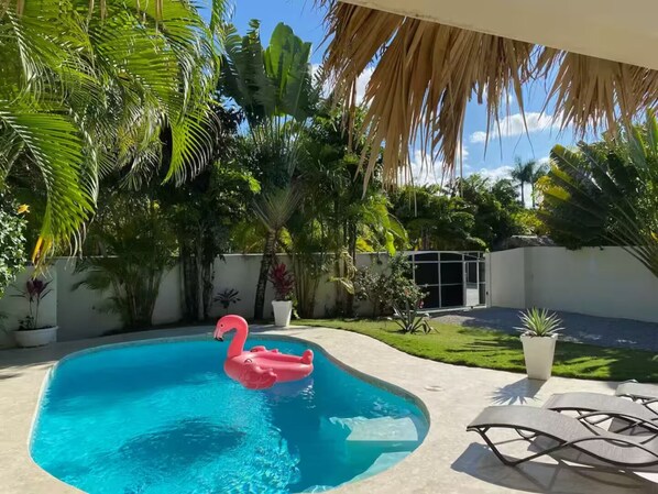 Ein Swimmingpool mit einem rosa Flamingo-Schwimmpunkt, umgeben von üppigen tropischen Pflanzen und einer weißen Wand.