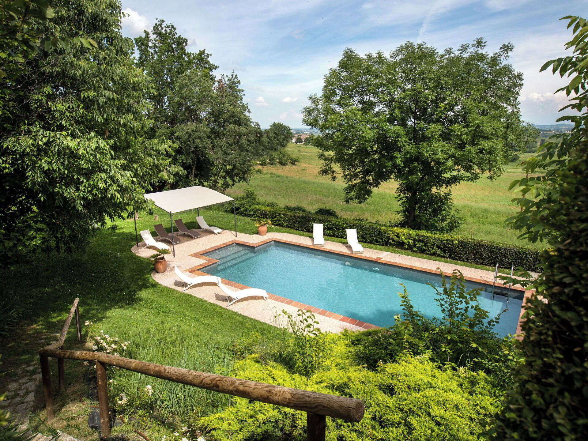 A rectangular swimming pool with a tiled deck, surrounded by lush greenery and trees.