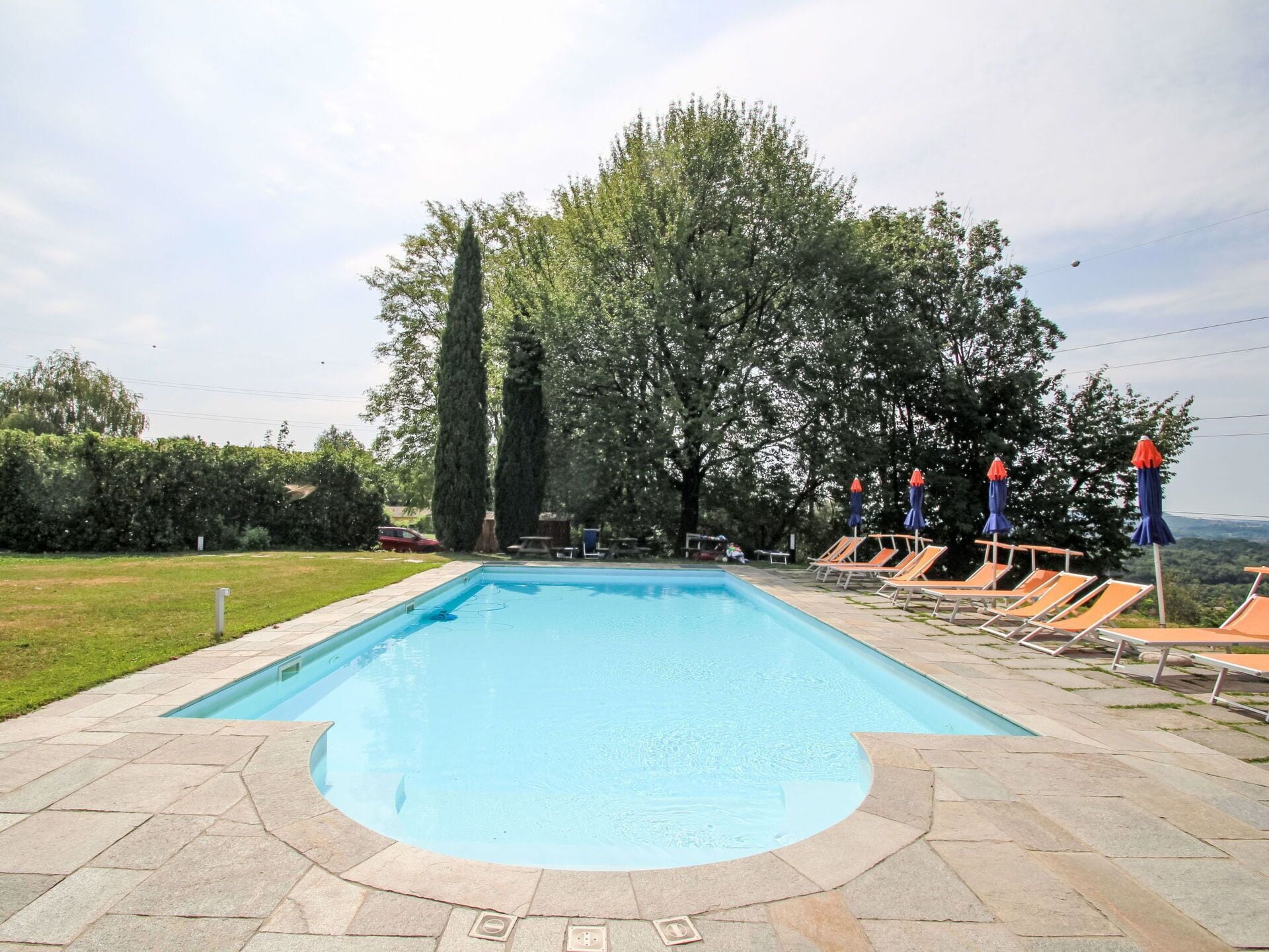 Ein rechteckiger Pool mit Steinrand, umgeben von Liegestühlen und Sonnenschirmen, mit Bäumen und blauem Himmel im Hintergrund.