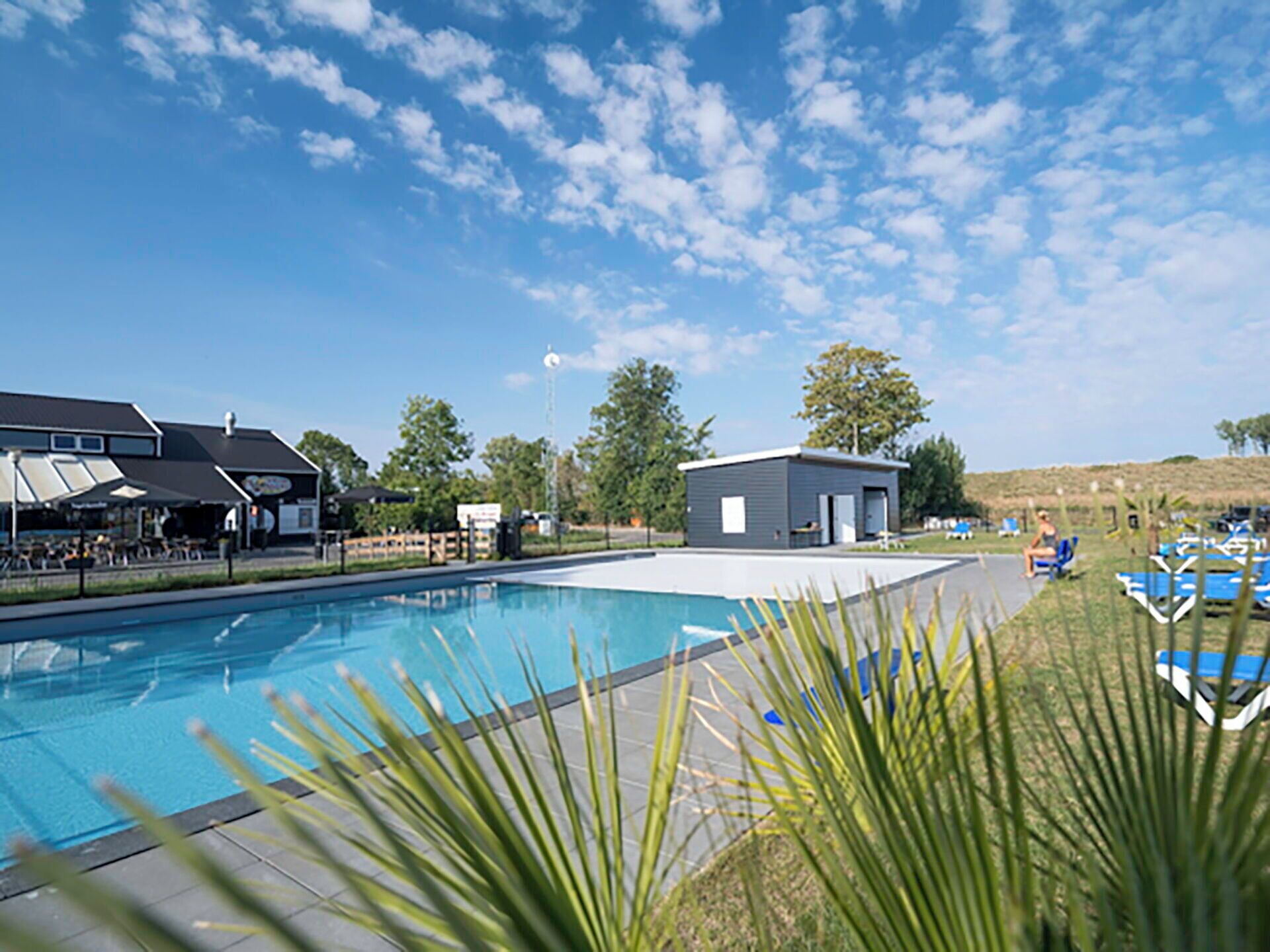 Une piscine avec des chaises longues et un petit bâtiment, le tout entouré de verdure sous un ciel dégagé.