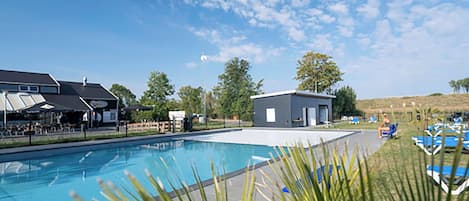 Une piscine avec des chaises longues et un petit bâtiment, le tout entouré de verdure sous un ciel dégagé.
