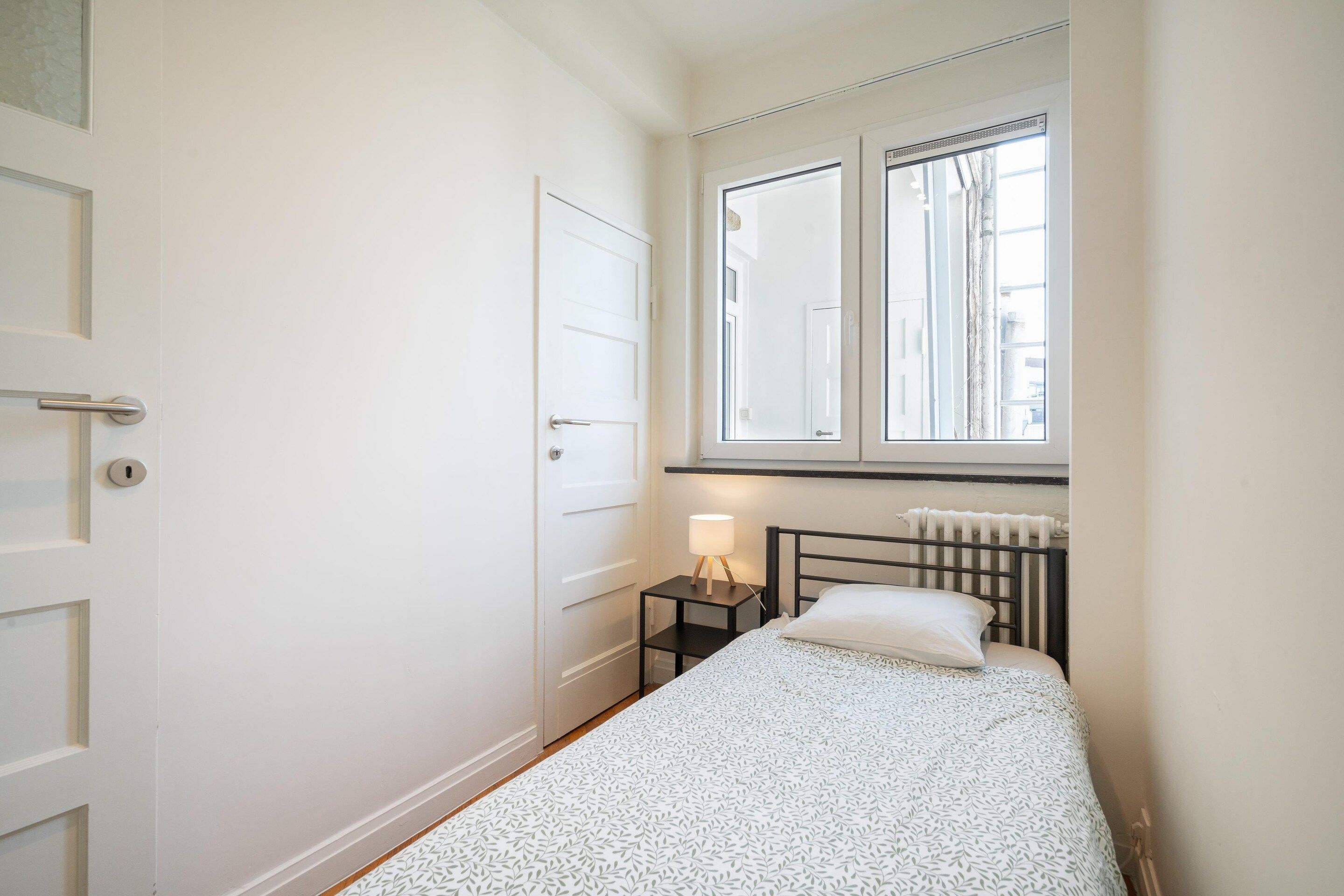 A small, single-bed room with a window, a radiator, and a bedside table with a lamp.