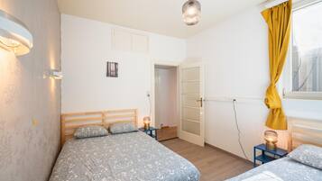 A bedroom with two beds, a door, a window with yellow curtains, and a hanging light fixture.
