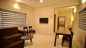 A hotel room with a sofa, a small round table, a dining table with chairs, a television mounted on the wall, and a ceiling fan. - Cocostay Kozhikode (Kozhikode)