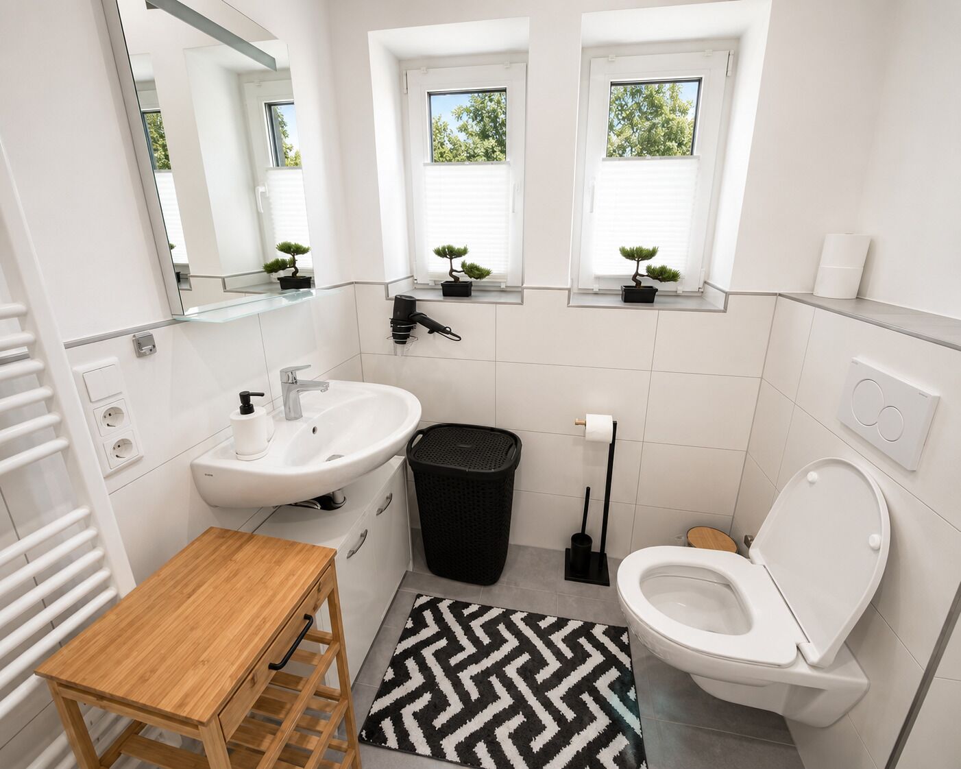 A modern bathroom with a white sink, toilet, and a small wooden table. There are three windows with plants on the windowsill, and a black trash bin is visible.