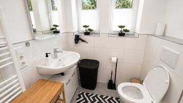 A modern bathroom with a white sink, toilet, and a small wooden table. There are three windows with plants on the windowsill, and a black trash bin is visible.