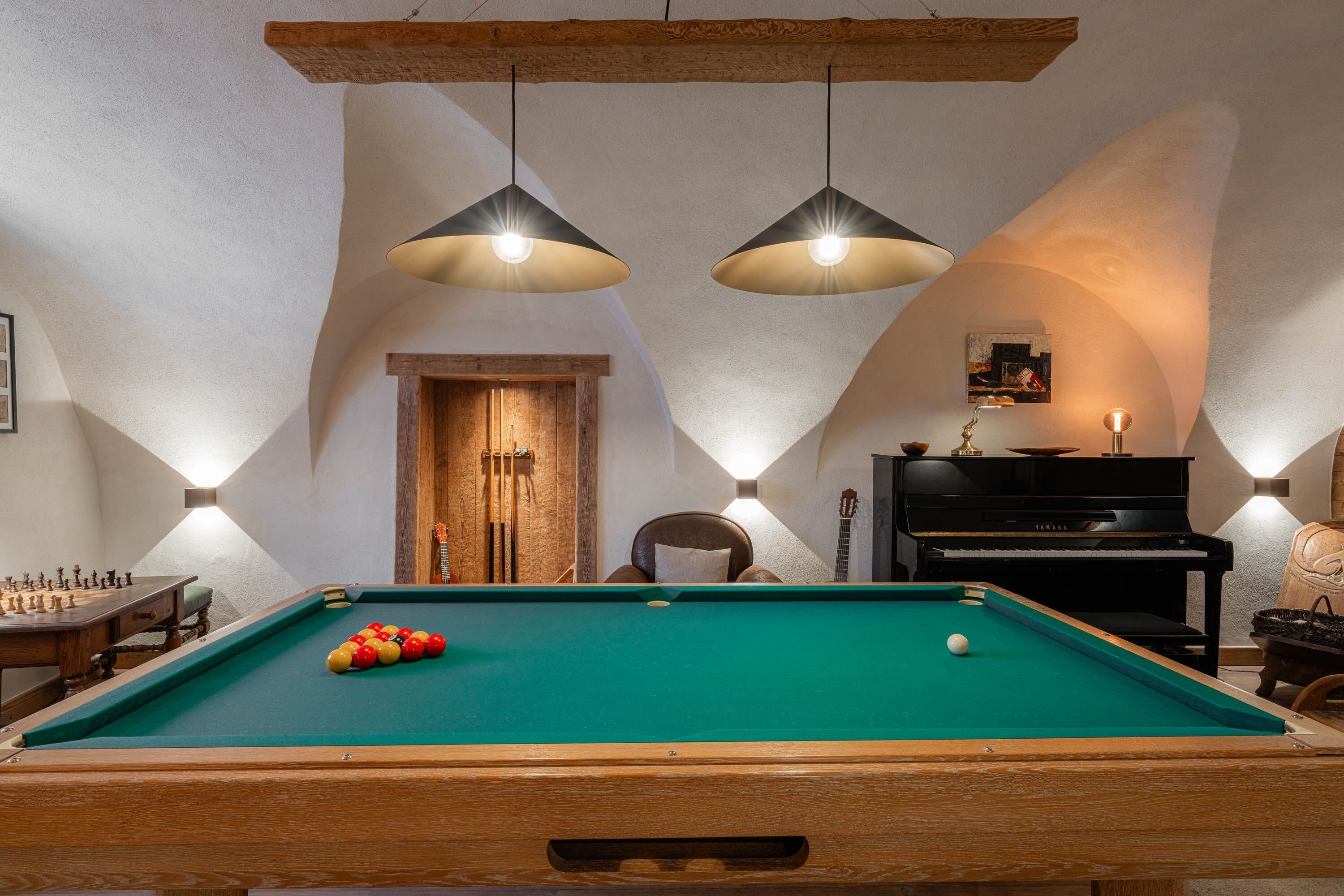 Una mesa de billar con superficie de fieltro verde, una silla de madera, un piano negro y dos lámparas colgantes.