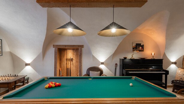 A pool table with a green felt surface, a wooden chair, a black piano, and two pendant lights.