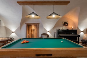 A pool table with a green felt surface, a wooden chair, a black piano, and two pendant lights.
