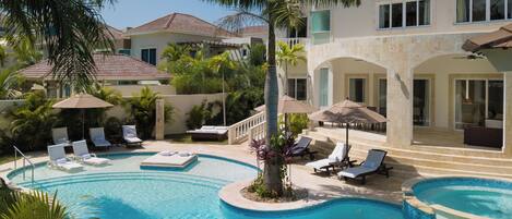 A resort pool area with multiple lounge chairs and umbrellas, surrounded by palm trees and a large building with a tiled roof.