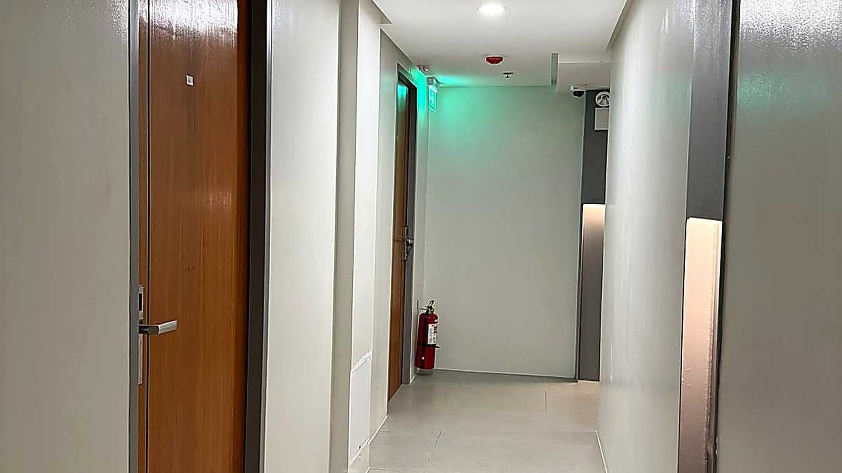 Corredor com uma porta de madeira à esquerda, um extintor de incêndio à direita e uma luz verde no final.