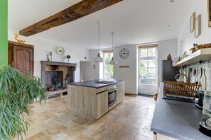 A spacious kitchen with a central island, wooden beams, a fireplace, and a large window. - Les Marquetons - Maison XIXe siècle pour 10 pers (Jaligny-sur-Besbre)