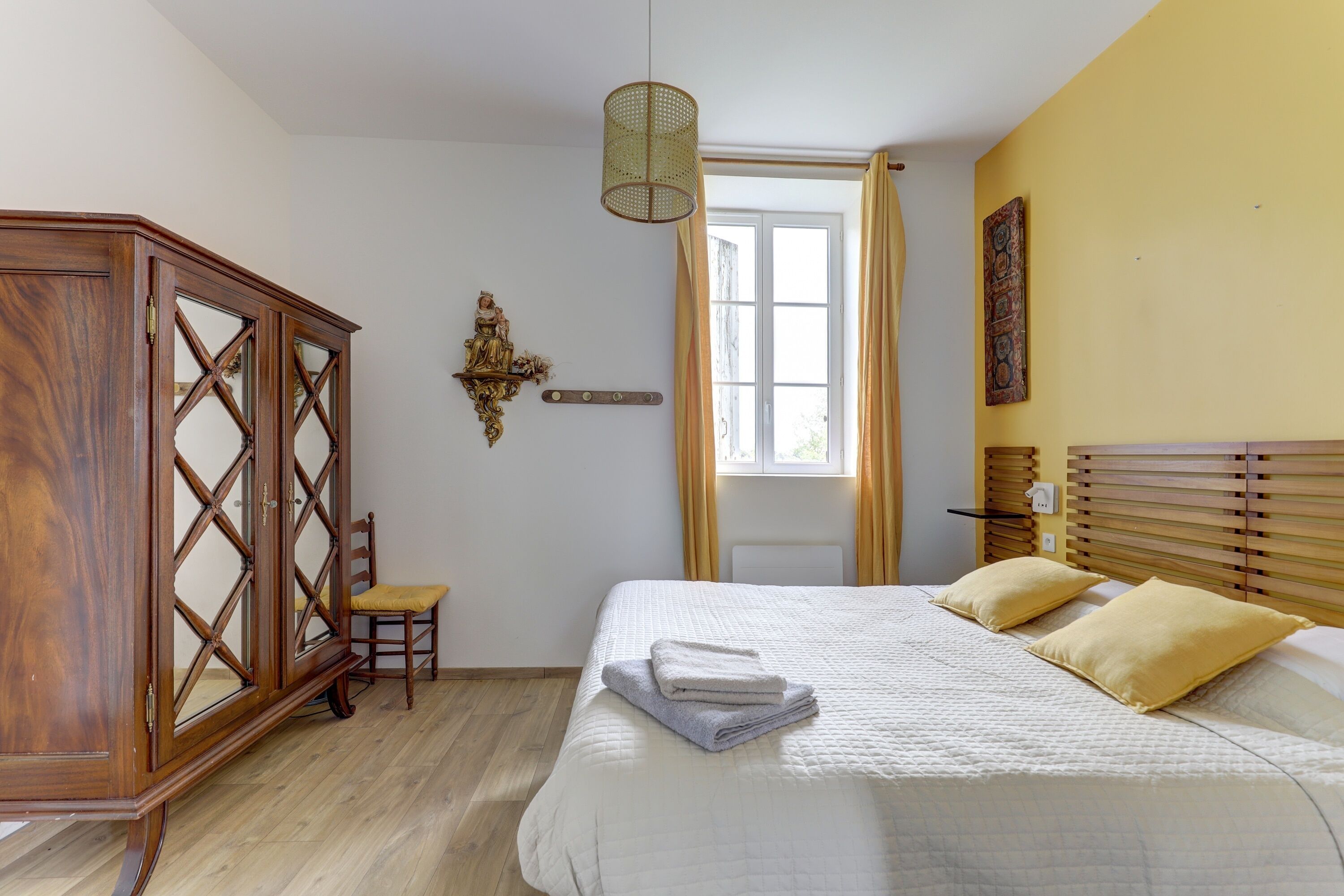 Un dormitorio con cama, un armario de madera, una silla, una ventana con cortinas y un aplique de pared.