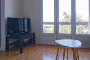 A room with a flat-screen TV on a stand, a small table, and a balcony with a railing. - La Bisquine Granvillaise - Appt 1 chambre proche de la plage (Granville)