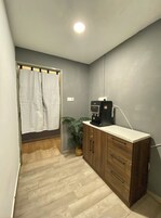 Una habitación pequeña y bien iluminada con un armario de madera, un dispensador de agua, una planta en maceta y una cortina de ducha.